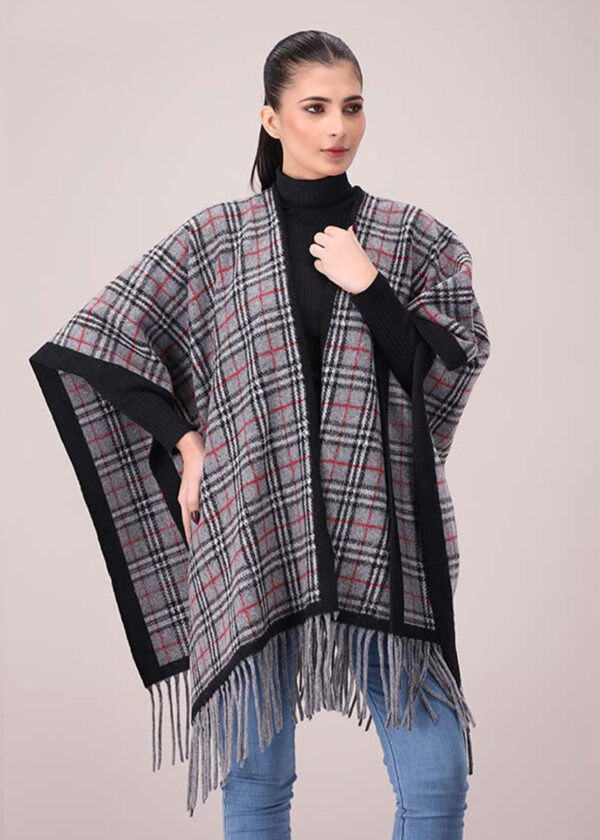 A woman posing, wearing a grey plaid open-front poncho with black and subtle red lines, and grey fringe along the bottom. She is wearing a black turtleneck and blue jeans.
