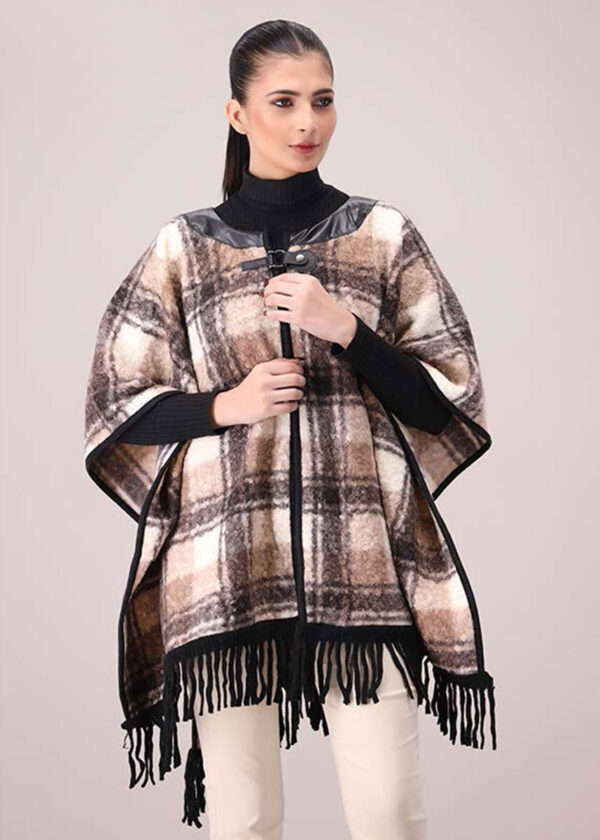 A woman wearing a brown, beige, and off-white plaid poncho with black leather-look trim around the neckline and down the front, featuring black fringe along the bottom. She is wearing a black turtleneck and light-colored pants.
